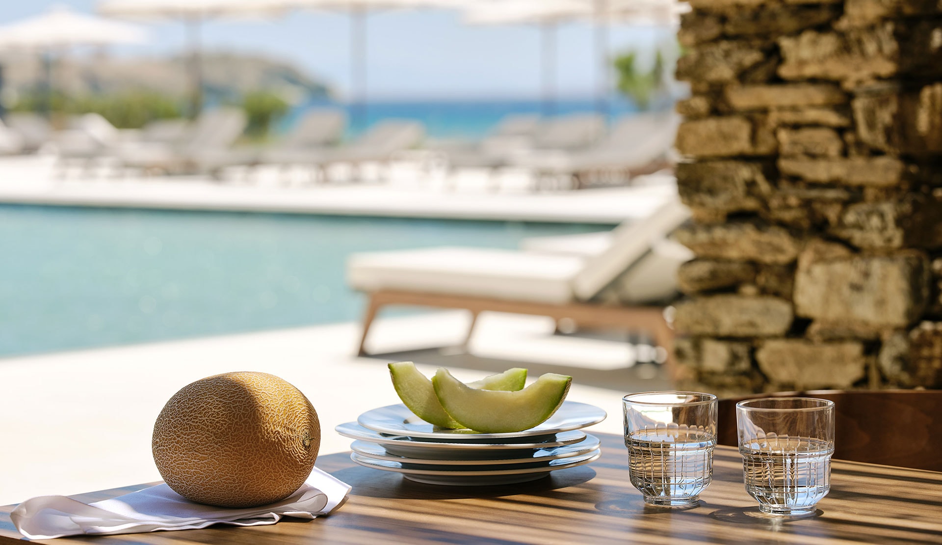 Melon and glasses of water on a table beside a pool and a stone wall